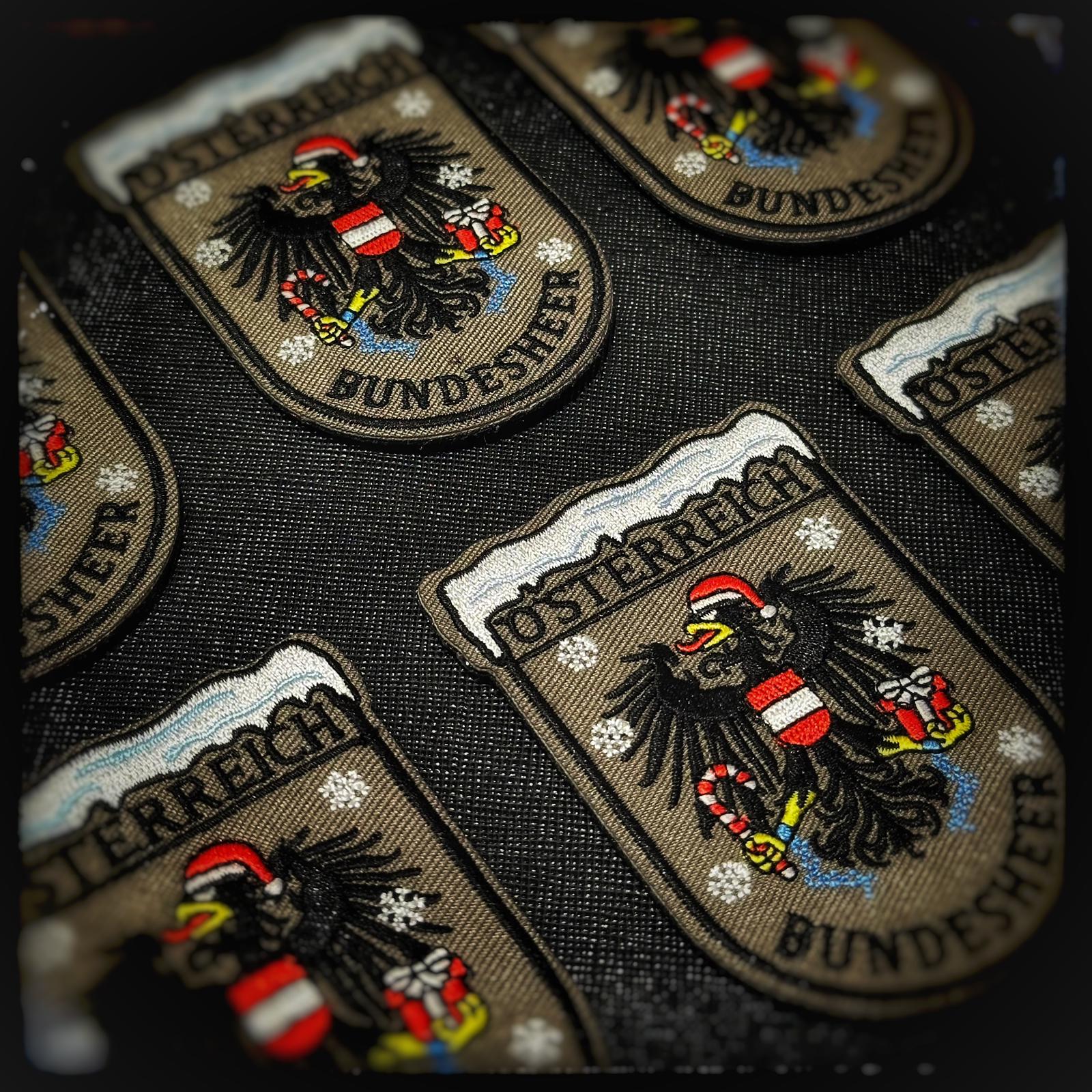 Gewebter Patch mit Klett: Bundesheer Weihnachtsadler. Österreichs stolzestes Wappentier zeigt sich weihnachtlich: der Bundesheer-Adler mit Nikolausmütze, Geschenk in der einen Kralle und Zuckerstange wie eine festliche Seitenwaffe in der anderen. Wachsam wie immer – nur kurz im Einsatz zum Schutz von Weihnachten. Hochwertige und langlebige 3D PVC, gewebte und gestickte Patches für Sammler, Airsofter und Militärangehörige. Dient auch als Moral-Patch. Patchworld.net. patchworld