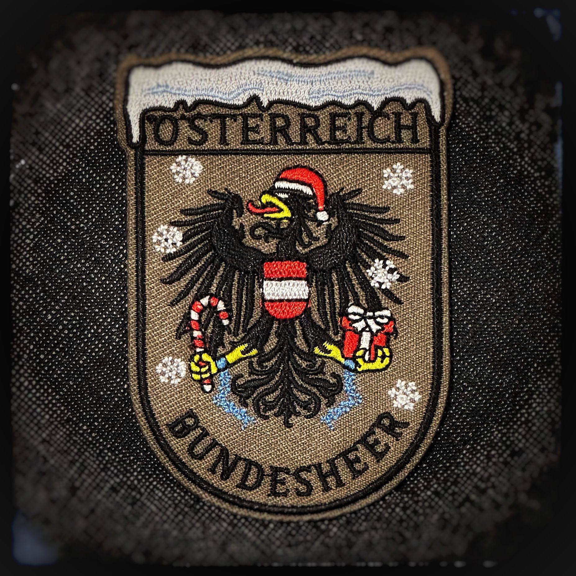 Gewebter Patch mit Klett: Bundesheer Weihnachtsadler. Österreichs stolzestes Wappentier zeigt sich weihnachtlich: der Bundesheer-Adler mit Nikolausmütze, Geschenk in der einen Kralle und Zuckerstange wie eine festliche Seitenwaffe in der anderen. Wachsam wie immer – nur kurz im Einsatz zum Schutz von Weihnachten. Hochwertige und langlebige 3D PVC, gewebte und gestickte Patches für Sammler, Airsofter und Militärangehörige. Dient auch als Moral-Patch. Patchworld.net. patchworld